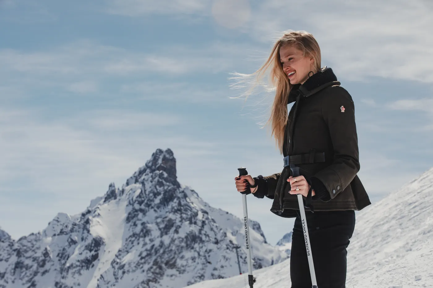 Woman wearing black Moncler ski wear posing with ski poles against the snowy mountains of Courchevel.