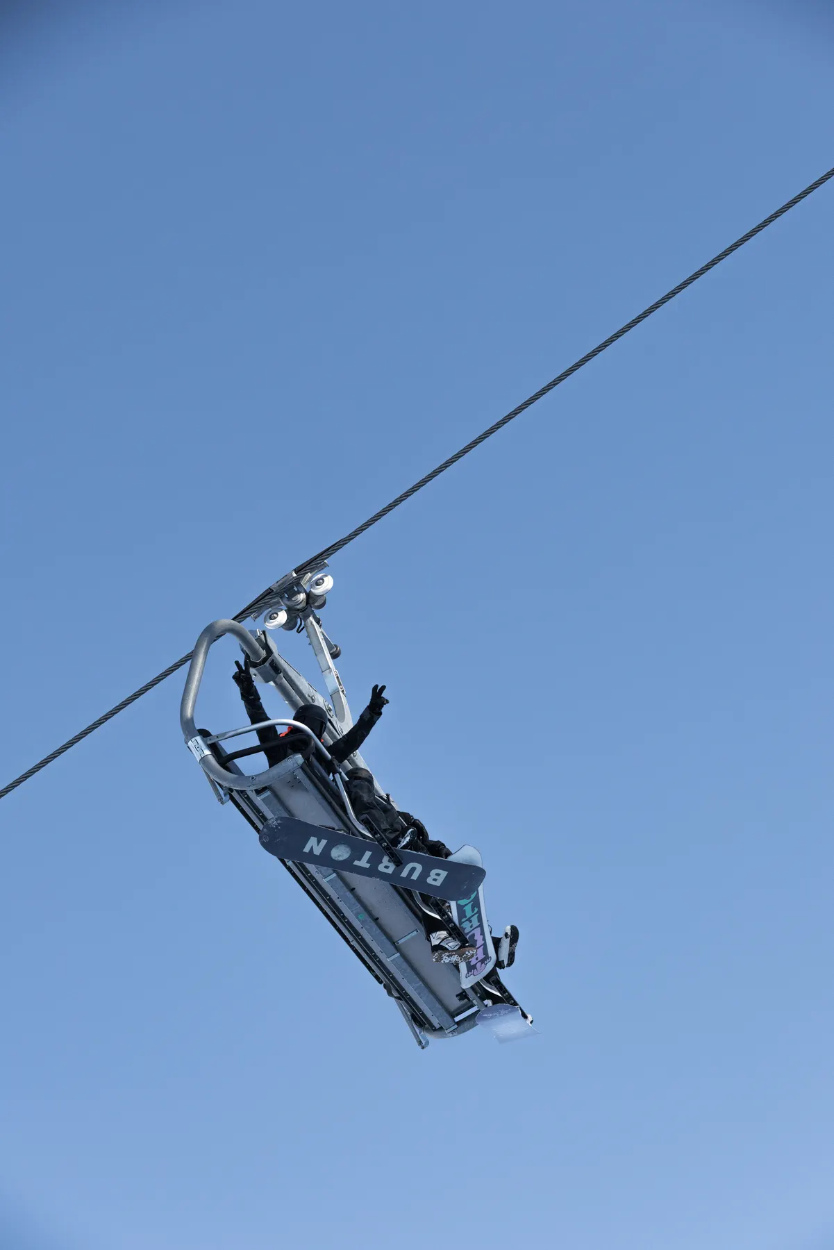Snowboarder riding a ski lift with a Burton snowboard against a clear blue sky in Courchevel.